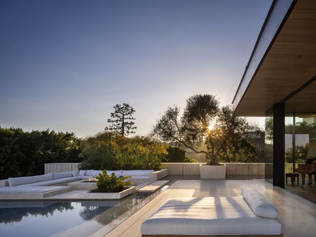 A sophisticated outdoor terrace beside a swimming pool, featuring built-in white seating, a raised planter, pale stone or concrete paving, and a covered indoor-outdoor living area with a deep timber-lined soffit and dark structural columns. Large glazed openings reveal a dining area inside. The setting is landscaped and viewed in warm late-afternoon light, with clean horizontal lines and a strong connection between the house and pool.