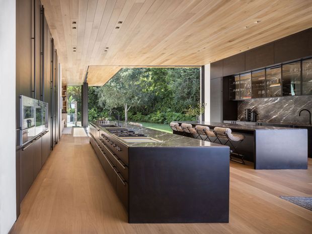A spacious, refined contemporary kitchen with a long central island, dark integrated cabinetry, a dramatic marble backsplash, and a timber-lined ceiling. Full-height glazing opens the room toward a lush garden, while warm timber flooring and ceiling boards create a cohesive, natural feel. The kitchen includes extensive built-in appliances, a cooktop, sink, bar seating, and a secondary dark island or peninsula.