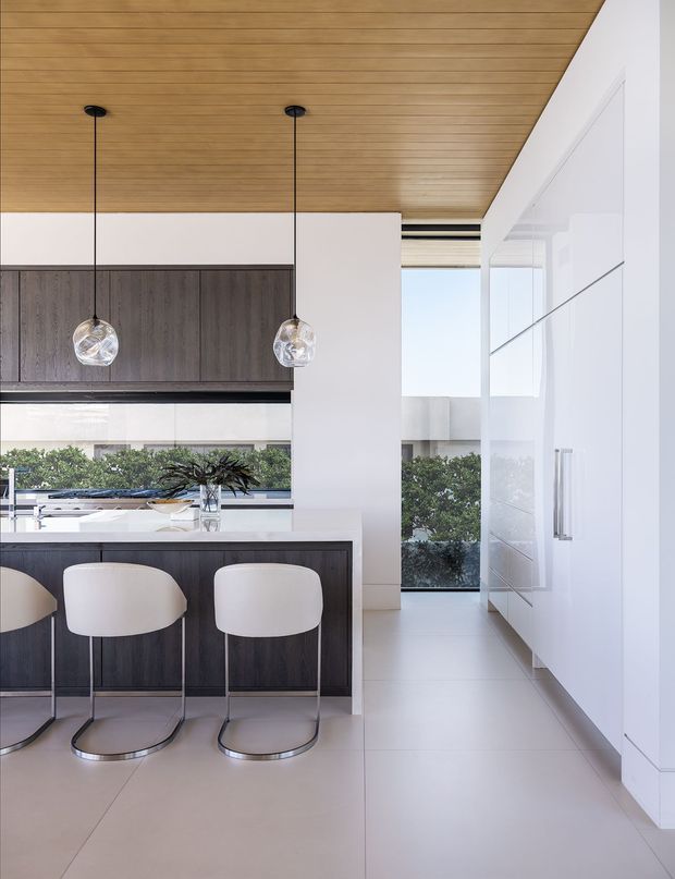 Bright contemporary kitchen with a central island and two white upholstered bar stools, dark wood-toned cabinetry, white full-height cabinetry, and a white benchtop. Clear glass pendant lights hang beneath a warm timber-lined ceiling. Large glazing and a reflective glass door bring in abundant daylight and overlook landscaped greenery.