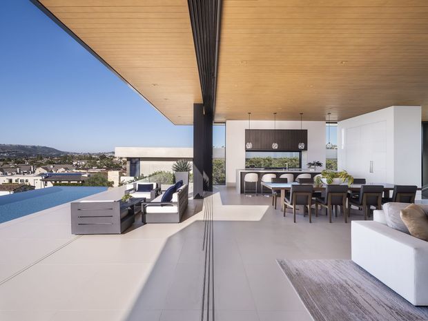 A spacious contemporary covered rooftop or terrace entertaining area with an infinity-edge pool overlooking a suburban hillside. The open-plan space combines an outdoor lounge, dining area, and fully fitted kitchen beneath a broad timber-lined soffit. Large pale floor tiles, white cabinetry, dark structural columns, and expansive open glazing create a bright indoor-outdoor setting.