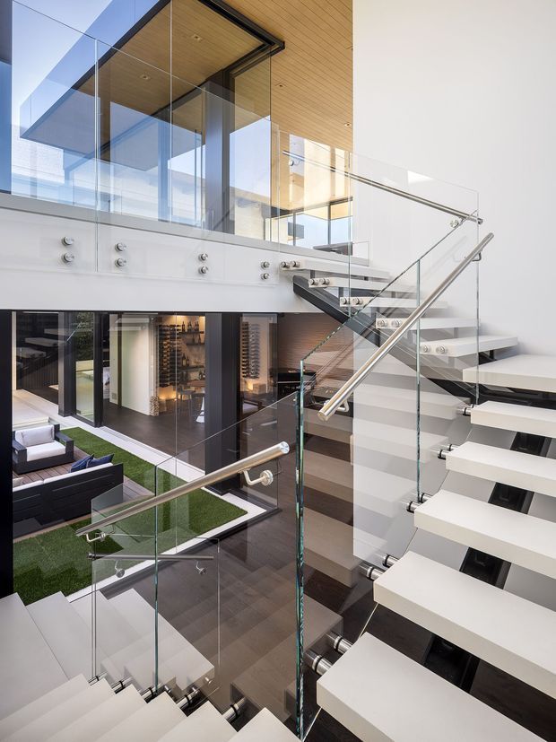 A dramatic multi-level contemporary interior focused on an open stairway. White floating stair treads with polished metal supports and handrails are enclosed by clear glass balustrades. The stair overlooks a lower living area with dark timber flooring, green rug-like flooring, sofas, and a visible wine display. The upper level features warm timber ceiling panelling, extensive glazing, and crisp white walls, creating a bright, refined architectural composition.