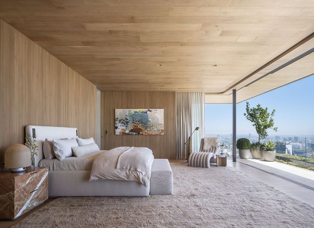 A luxurious contemporary bedroom with a low-profile upholstered bed, soft neutral bedding, a large textured rug, and warm vertical timber wall and ceiling lining. Full-height sliding glass doors open to a terrace with potted plants and expansive city views. A sculptural lounge chair, bedside furniture, artwork, and layered neutral textures create a calm, refined atmosphere.