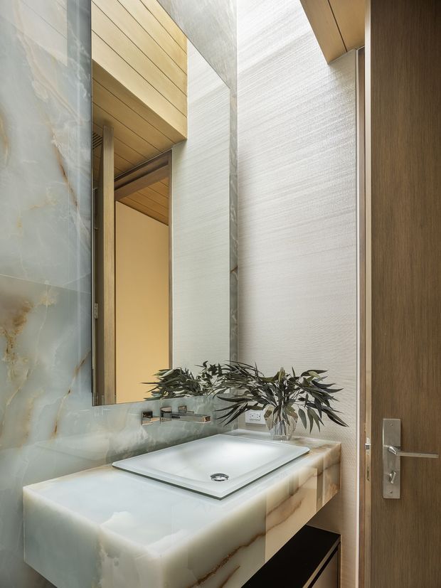 A refined contemporary bathroom vanity viewed beside an open timber door. The room features a large pale translucent stone vanity and integrated basin, a matching pale stone backsplash, a wall-to-wall mirror, warm timber ceiling lining, and a dark timber door. Natural foliage in a glass vase adds an organic accent.