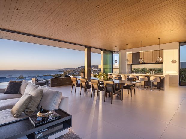 A spacious open-plan coastal living, dining and kitchen interior opening onto a broad terrace with expansive ocean views. The room features a warm timber-lined ceiling, full-height glazing, pale large-format flooring, a contemporary kitchen with an island and bar seating, a dining table, and a large sectional sofa. The design is bright, refined and strongly connected to the landscape.