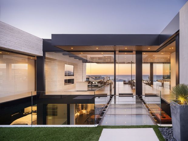A contemporary multi-level home organized around a glazed central courtyard and transparent bridge. Dark charcoal cladding and slim black-framed glazing contrast with pale textured walls and illuminated interiors. The view looks through the house toward a coastal horizon, with a rooftop lawn, pale stepping path, gravel planting bed, and sculptural yucca visible in the foreground. Interior areas include a living space, dining area, and lower-level lounge visible through the glazing.
