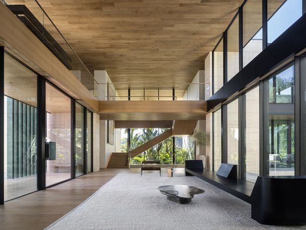 A spacious, refined double-height living room or gallery-like lounge with extensive floor-to-ceiling glazing, warm timber ceilings and floors, pale stone or plaster walls, and black-framed windows and doors. A mezzanine bridge crosses the room, with a visible staircase and abundant greenery outside. Minimal dark seating and a sculptural low coffee table sit on a large pale rug.