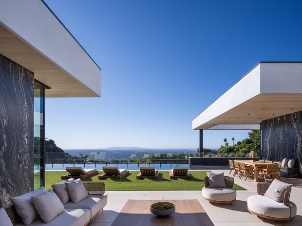 A luxurious modern outdoor terrace and lounge between two flat-roofed white pavilions, overlooking a swimming pool and expansive distant landscape. The space features pale stone or concrete paving, timber-toned soffits, dark veined stone feature walls, outdoor sofas and lounge chairs, a dining setting, manicured lawn, and floor-to-ceiling glazing.