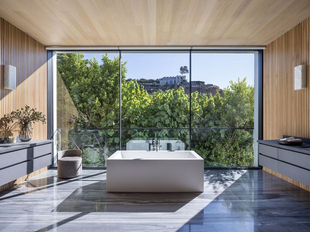 A refined contemporary bathroom with a freestanding white bathtub centred before expansive floor-to-ceiling glazing. Warm vertical timber wall slats and a matching timber ceiling contrast with dark grey cabinetry and a polished dark stone floor. The room is filled with natural light and overlooks dense greenery and a rocky hillside.