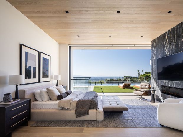Bright, refined contemporary living room with a large sectional sofa, upholstered ottoman, armchair, wall-mounted television and fireplace. The room features a light timber-lined ceiling, pale timber flooring, a dramatic dark stone or marble fireplace wall, framed artwork, and expansive glazed doors opening to a landscaped terrace with ocean views.