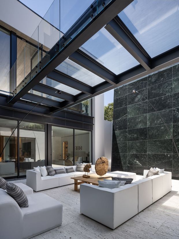 A dramatic contemporary outdoor courtyard lounge enclosed by glazed walls and a large steel-framed glass canopy. White sectional sofas and low timber coffee tables are arranged on a pale stone or concrete floor, with a tall dark green stone feature wall and black structural framing creating strong contrast. The space is bright, refined, and open to the sky through transparent glazing.