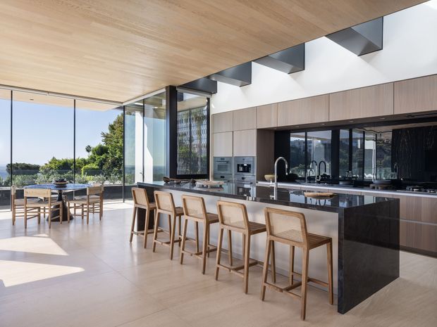 A spacious, refined contemporary kitchen and dining area with a long island, integrated dark stone sides, timber cabinetry, and several woven-seat timber bar stools. Full-height glazing opens to a leafy waterfront or landscaped outlook, while a timber-lined ceiling and clerestory-like roof glazing bring in abundant natural light.