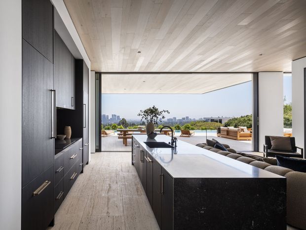 A sophisticated open-plan kitchen with a large dark island and integrated sink, set beneath a light timber-lined ceiling. Floor-to-ceiling sliding glazing opens directly to an outdoor entertaining terrace and pool, with a city skyline visible beyond. Dark built-in cabinetry contrasts with pale timber flooring and ceiling, while a living area is partially visible to the right.