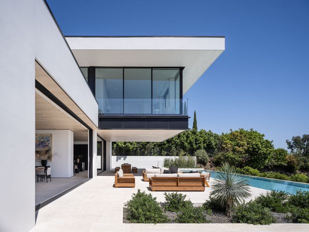 Contemporary multi-level residential exterior with crisp white rendered walls, expansive black-framed glazing, cantilevered upper-floor geometry, and warm timber soffits. The view looks onto a spacious paved terrace furnished with outdoor seating, landscaped planting, and a long swimming pool beside the house.