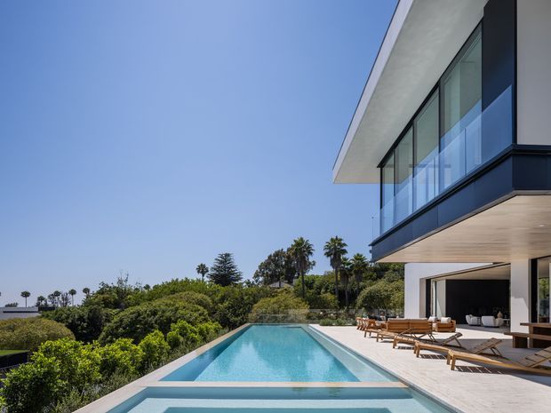 A contemporary multi-level residence viewed from the pool terrace, featuring a long infinity-edge swimming pool, pale stone or concrete paving, extensive floor-to-ceiling glazing, a projecting upper floor with a glass balustrade, and a clean white and dark charcoal facade. Outdoor lounge furniture sits beside the open-plan ground floor, with landscaped greenery and palms beyond.