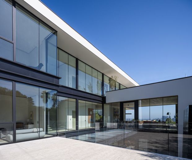 Contemporary two-storey residential house with crisp white flat roof planes, extensive floor-to-ceiling glazing, dark slender window frames, and a sheltered central courtyard or terrace. The glass-lined outdoor area overlooks a distant coastal or urban horizon, while a minimalist living space is visible through the glazing.
