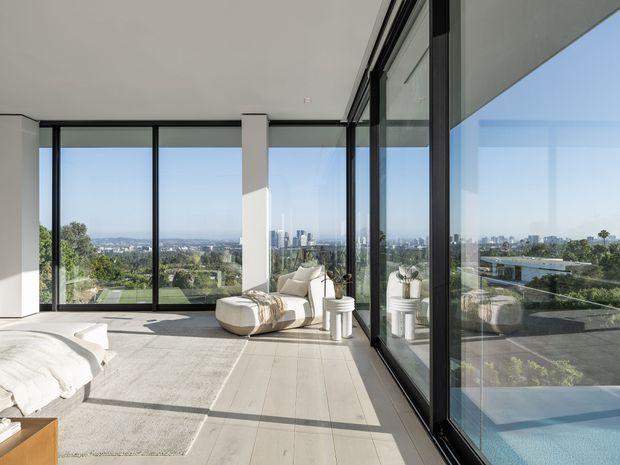A bright, contemporary bedroom or sitting area with expansive floor-to-ceiling black-framed glazing overlooking a city skyline and landscaped surroundings. The room has pale stone or concrete-look flooring, white walls, a low white bed or seating at left, and a sculptural cream lounge chair beside a small round side table. Strong sunlight and reflections create an airy, refined atmosphere, with a blue pool or water feature visible outside through the glazing.