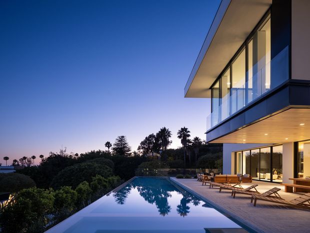Twilight exterior view of a sleek multi-level modern residence with expansive floor-to-ceiling glazing, projecting flat roof planes, glass balcony balustrades, and warm recessed lighting. A long infinity-edge swimming pool and timber-toned pool deck occupy the foreground, with landscaped greenery and palm trees beyond.