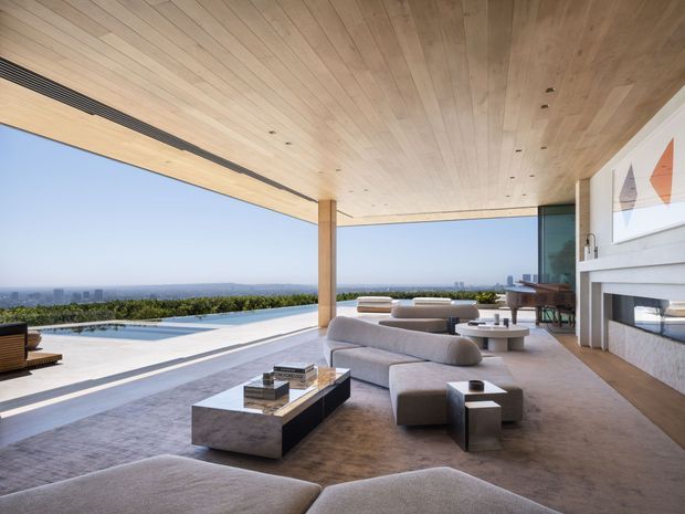 A luxurious open-plan living room extending onto a terrace and pool, with expansive panoramic glazing and an unobstructed horizon view. The space features a warm timber-lined ceiling with recessed lighting, pale neutral upholstered sectional seating, low stone coffee tables, a built-in linear fireplace, and a dining area beyond. The architecture is strongly indoor-outdoor, with slender timber posts, broad roof overhangs, and clean contemporary lines.