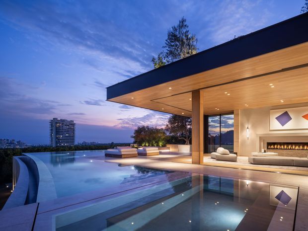 Twilight view of a luxurious modern residence with a broad cantilevered flat roof, warm timber-lined soffit, exposed structural columns, expansive glazing, and an open-plan living area with a fireplace. An infinity-edge swimming pool and reflective water feature dominate the foreground, with a landscaped terrace and distant urban skyline beyond.
