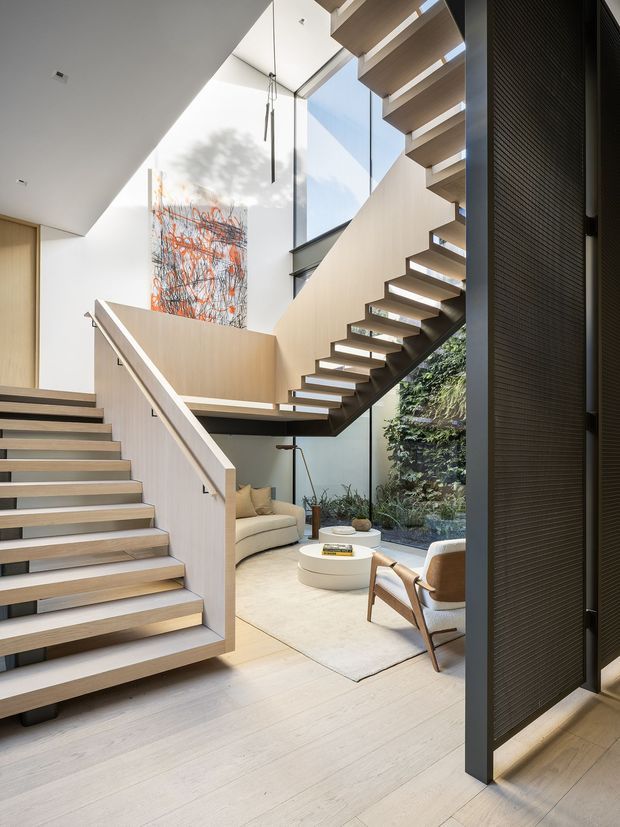 A bright contemporary interior focused on a sculptural open-riser staircase spanning multiple levels. Pale timber stair treads, light wall panels and flooring contrast with dark structural elements and a tall black-framed glazed wall. A lower-level living area with a cream sofa, round coffee tables, armchair and indoor planted wall is visible beneath the stair, with a large abstract artwork and suspended linear pendant lights adding visual interest.