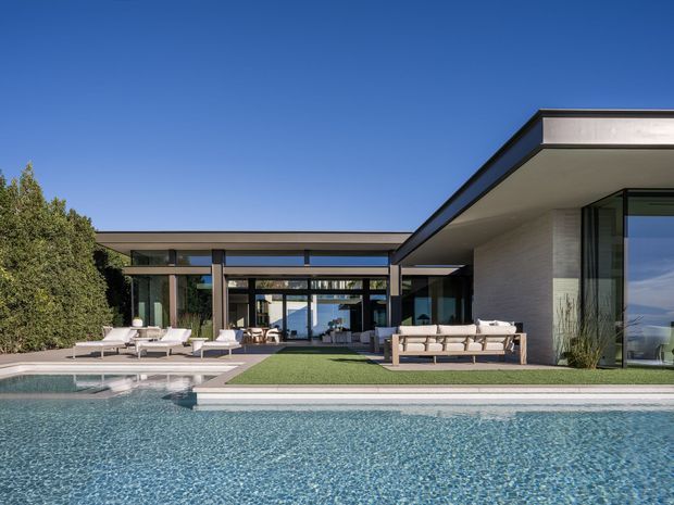 Exterior view of a large contemporary single-storey residence arranged around a swimming pool. The house features strong horizontal roof planes, extensive floor-to-ceiling glazing, dark slender frames, pale masonry walls, and a covered indoor-outdoor entertaining zone with outdoor seating and lounge chairs. The landscaped setting includes clipped hedges and a broad pool terrace.