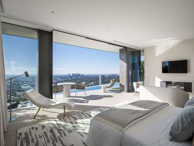 Bright, luxurious high-rise bedroom with a large bed in the foreground, floor-to-ceiling glazing, and expansive city and water views. The room has pale timber flooring, a light neutral rug, sculptural lounge seating, a small table, a wall-mounted television, and a sleek contemporary fireplace or built-in feature wall. Sliding glass panels open toward a balcony or terrace.