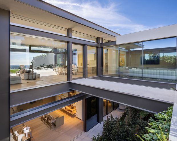 A contemporary multi-level residence organized around a landscaped internal courtyard. Floor-to-ceiling glazing, dark structural frames, cantilevered upper-level walkways, and open-plan living and kitchen areas create a bright, highly connected indoor-outdoor composition. A glimpse of coastal water is visible beyond the upper living area.