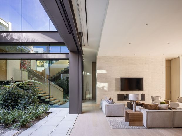A bright contemporary living room opening onto a landscaped internal courtyard, with extensive floor-to-ceiling glazing, a floating staircase with glass balustrade, pale timber flooring, and a textured light stone or masonry feature wall surrounding a wall-mounted television. Neutral upholstered seating and restrained furnishings create an airy, refined interior with strong indoor-outdoor connection.