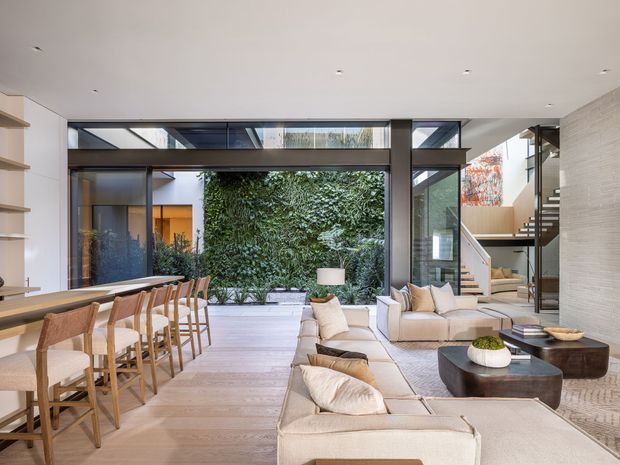 A bright, spacious contemporary open-plan living area with a dining zone and kitchen island in the foreground, a large upholstered sectional seating arrangement, and a pair of dark rounded coffee tables. Full-height glazed openings connect the room to a planted internal courtyard, while an open staircase with timber treads and a glass balustrade rises at the right. The interior uses pale timber flooring, light neutral upholstery, white walls and ceilings, and dark metal-framed glazing and structural elements.