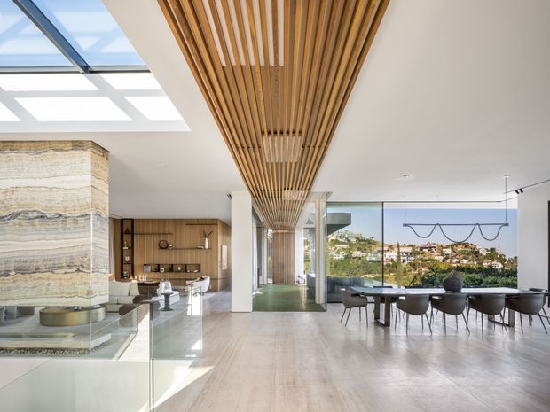 A bright, open-plan contemporary interior combining a living area, dining area and kitchen/bar zone. Floor-to-ceiling glazing overlooks a leafy suburban landscape, while a dramatic linear timber-slat ceiling feature runs through the space. A large layered natural-stone fireplace forms a sculptural focal point beside a glass balustrade. The room has pale stone flooring, white walls and columns, warm timber cabinetry, a dark dining table with modern chairs, and abundant natural light from skylights and glazing.