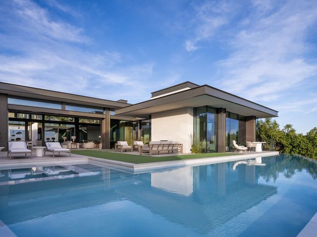 Exterior view of a large contemporary single-storey residence with broad flat roof planes, extensive floor-to-ceiling glazing, and an open-plan indoor-outdoor living area overlooking a swimming pool. The pale rendered facade, dark metal-framed glazing, covered terrace, outdoor seating, and reflective pool create a clean, resort-like composition.