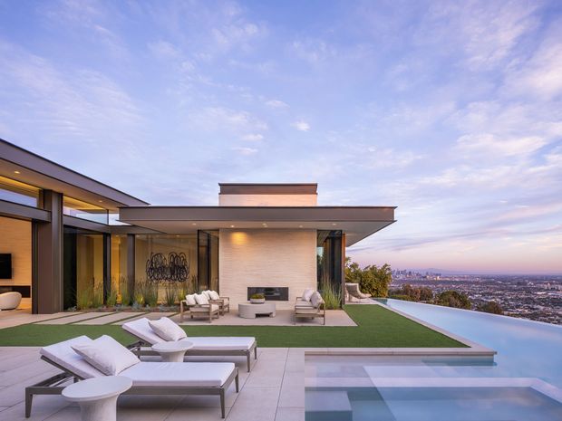 A luxurious modern rooftop or elevated terrace with a flat-roofed residence, expansive glazing, an outdoor living area, fireplace feature wall, artificial lawn, sun loungers, and an infinity-edge swimming pool overlooking a broad urban landscape. The composition is contemporary and highly open, with clean horizontal lines and a pastel evening sky.