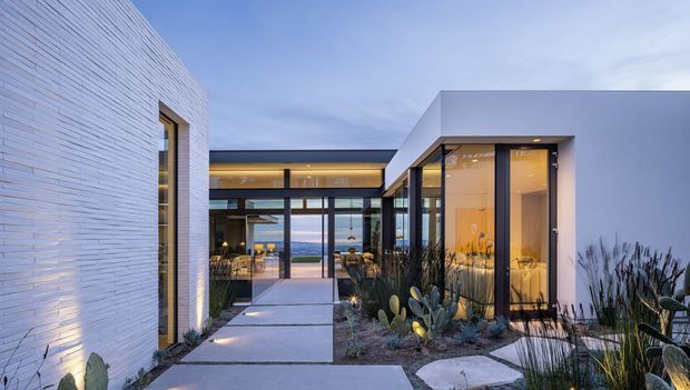 Contemporary single-storey residence with crisp white rendered and finely textured masonry walls, extensive black-framed glazing, and low flat roof forms arranged around a landscaped courtyard walkway. Warm interior lighting glows through the glass, while drought-tolerant grasses and sculptural cacti frame the concrete stepping path. A distant blue landscape is visible through the central glazed living area.