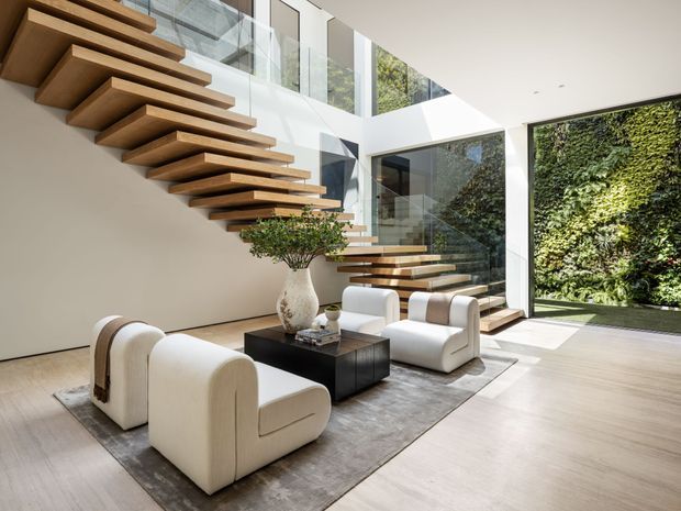 A bright, sophisticated double-height living area with dramatic floating timber staircases, expansive floor-to-ceiling glazing, and a lush vertical garden visible outside. Two cream boucle-style lounge chairs and matching sculptural seating surround a black coffee table on a large grey rug. A sculptural vase and greenery add an organic touch, while the pale timber floor and white walls create a clean, contemporary atmosphere.