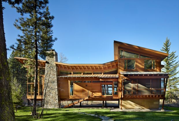 Exterior view of a multi-level timber-and-stone house set among mature trees. The building features extensive warm timber cladding and structural beams, a prominent natural-stone chimney, broad glazed openings, covered outdoor walkways, and a strongly articulated sloping roofline. A lawn and stepping-stone path lead toward the elevated entrance.