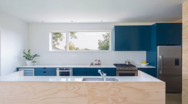 Bright contemporary kitchen with a large central island clad in pale plywood, white benchtops, and bold deep-blue cabinetry. The room includes a stainless-steel refrigerator, range cooker, dishwasher, sink, and wide horizontal windows overlooking trees. The clean-lined design has a light, airy feel with minimal decoration.