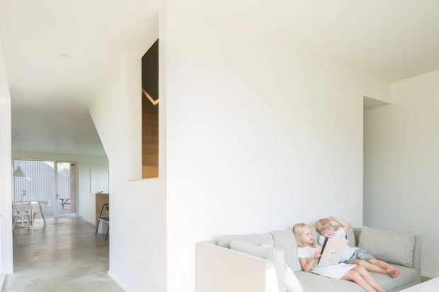 Bright minimalist open-plan interior with a white living room in the foreground, featuring a pale sectional sofa and children seated together. Through the opening is a light dining area with a timber dining table and chairs, pendant lighting, and full-height glazed doors. The space has white walls and ceilings, pale polished concrete flooring, and a simple recessed timber-and-dark feature niche.