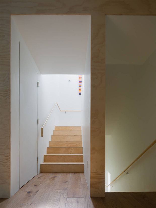 Interior view of a minimalist stairway with pale timber flooring, stair treads, wall panelling and door surrounds. White walls and a simple timber handrail lead up a compact flight of stairs, while a second stair opening is visible to the right. The space is bright, restrained and materially focused.