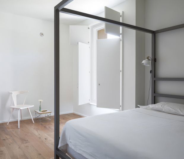 Bright, minimalist bedroom with a dark metal four-poster bed, white bedding, white walls, and pale timber flooring. A white chair and small side table sit near the wall, while open white doors lead to an adjacent area. The room has a clean, understated contemporary aesthetic with strong black-and-white contrast.