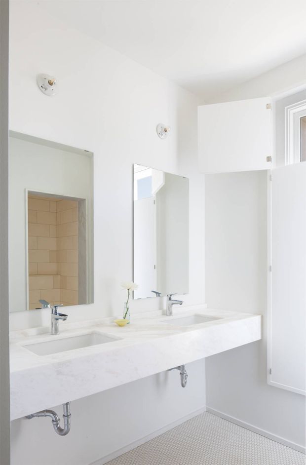 Bright, minimalist bathroom with a long double-sink vanity, white marble-look benchtop, polished chrome faucets and exposed plumbing. Large wall mirrors reflect the vanity and a tiled shower recess with warm beige rectangular tiles. White walls, a white ceiling, and small light mosaic floor tiles create a clean, airy interior.