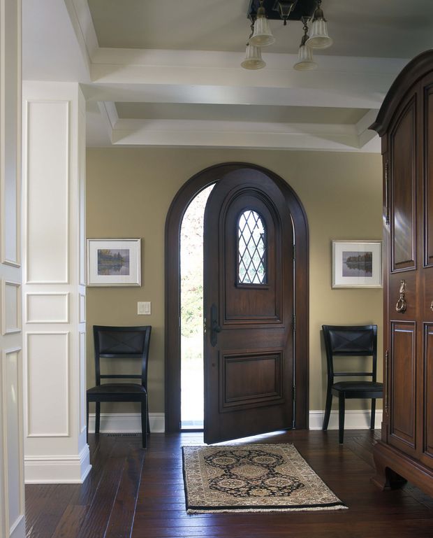 A formal residential entryway with a richly stained arched timber front door featuring decorative paneling and a diamond-leaded glass inset. The entry has dark timber flooring, pale olive-beige walls, white raised wall paneling and coffered ceiling details, traditional wall art, black leather chairs, a patterned runner rug, and a large dark timber built-in cabinet. A small multi-arm pendant light hangs overhead.