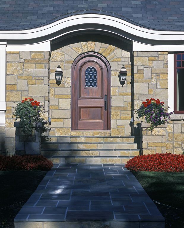 Front exterior entry of a formal stone-clad house, centred on a dark timber arched door with diamond-pattern glazing and decorative lantern sconces. A strongly curved, layered roof or canopy line frames the doorway, while broad stone steps lead to a slate-paved path bordered by manicured planting and flower planters.
