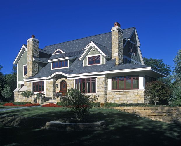 Exterior photograph of a large multi-storey detached house with steep intersecting gable roofs, dark slate-like roofing, pale stone veneer walls, green-grey painted siding, prominent stone chimneys, and red-brown framed windows. The landscaped front yard includes stone retaining walls and garden beds. The architecture has a strongly traditional, formal character with substantial masonry detailing.