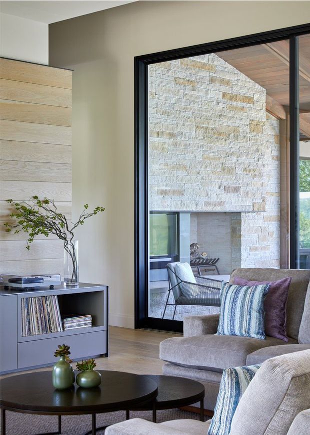 A contemporary living room with a neutral palette, built-in light timber wall panelling, low grey media/storage cabinetry, and a pair of dark round coffee tables. A large black-framed glazed opening provides views into a stone-lined courtyard or exterior seating area. Soft grey upholstered seating and striped blue-grey cushions add texture, while sculptural greenery and records provide restrained decor.
