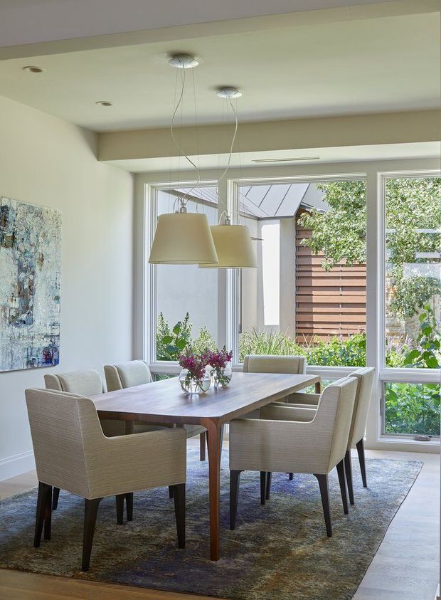 Bright contemporary dining room with a rectangular timber dining table surrounded by upholstered armchairs, a large area rug, paired pendant lights, recessed ceiling lighting, and expansive white-framed windows overlooking a landscaped courtyard. A large abstract artwork adds colour to the otherwise neutral interior.