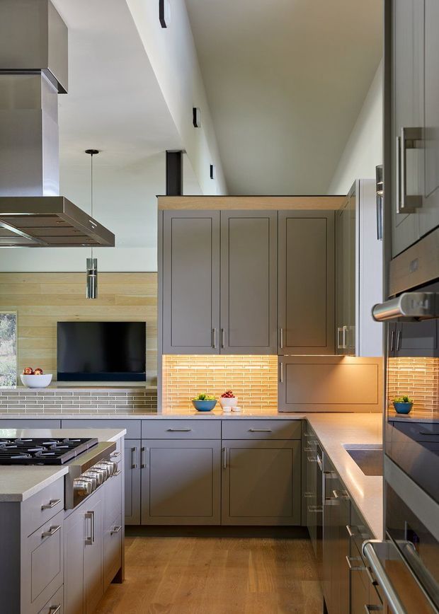 A refined contemporary kitchen viewed from within the room, featuring taupe-gray paneled cabinetry, pale stone benchtops, integrated stainless-steel appliances, a central island with gas cooktop, and warm timber flooring. A horizontal orange-toned brick backsplash with under-cabinet lighting adds texture and warmth, while a timber-clad wall and television are visible beyond the kitchen.