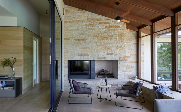 A refined mid-century-inspired living room with a substantial stacked natural-stone fireplace wall, warm timber-lined ceiling with exposed beams, dark-framed floor-to-ceiling glazing, and an open connection to a timber-finished hallway or entry area. Two lounge chairs and a small round table face the fireplace, while a built-in bench runs along the glazed wall. The room is bright and connected to greenery outside.