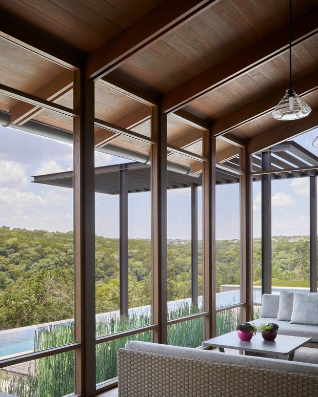 A covered, glass-enclosed outdoor lounge or sunroom with expansive views over a wooded landscape. The space features dark timber-framed glazing, a warm timber-lined ceiling with exposed beams, a hanging glass pendant light, a pale sectional sofa, woven lounge seating, and a low rectangular coffee table. A swimming pool and planted screening are visible beyond the glazing.