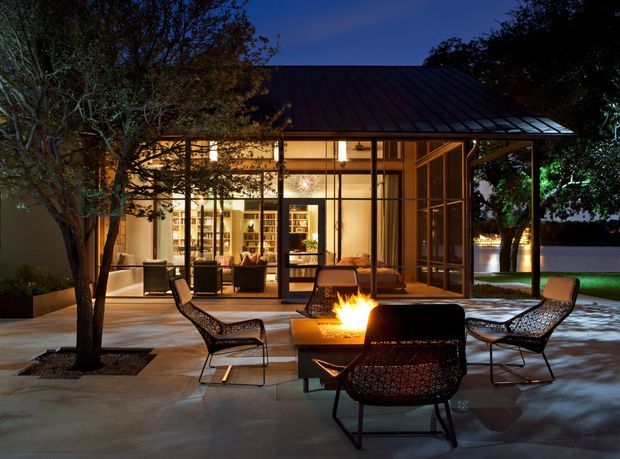 Twilight exterior view of a modern pavilion-like residence with extensive floor-to-ceiling glazing, a low pitched dark metal roof, and a warmly illuminated interior visible through the windows. A paved terrace in the foreground is furnished with woven lounge chairs around a square fire pit, with mature trees and landscaped planting surrounding the space.