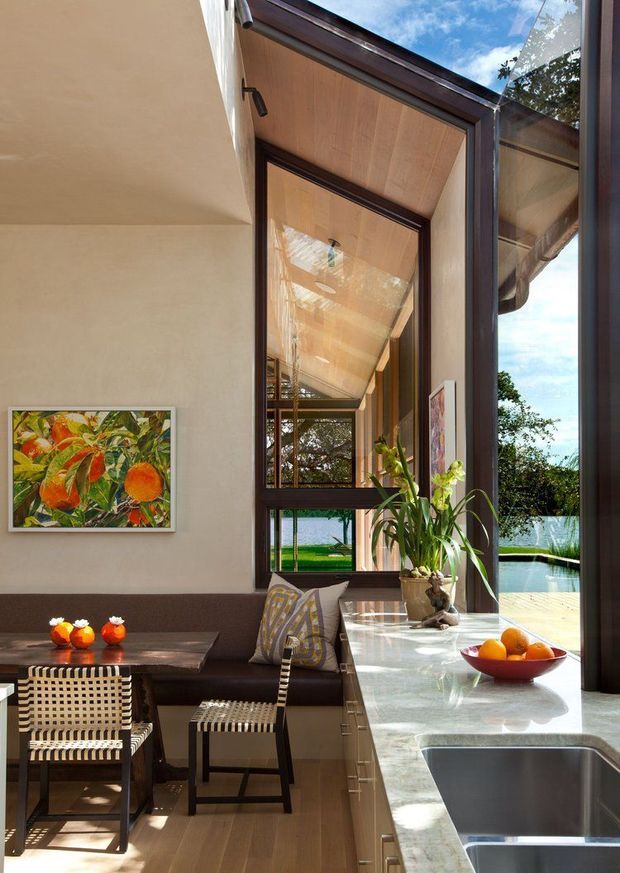 A warm, light-filled open-plan kitchen and dining area with a long pale stone island, integrated sink, dark cabinetry, and floor-to-ceiling dark-framed glazing. A built-in upholstered banquette and woven dining chairs sit beside a timber dining table. The glazing opens views to a landscaped pool and water beyond, while timber-lined ceiling surfaces and a large indoor plant add natural character.
